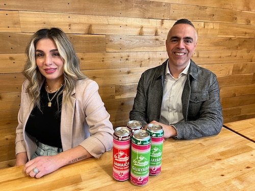 A woman and man sit at a wooden table in front of a wooden wall with green and red cans of cider in front of them.