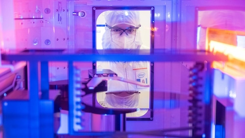 An Intel employee in a white bunnysuit looks into a manufacturing tool under bright factory lights.